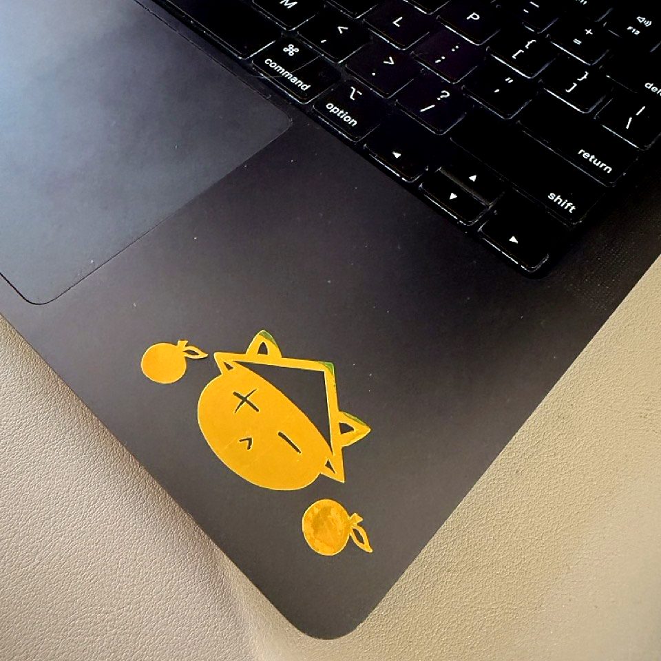A gold cat decal with two tangerines on the palm rest of a black MacBook