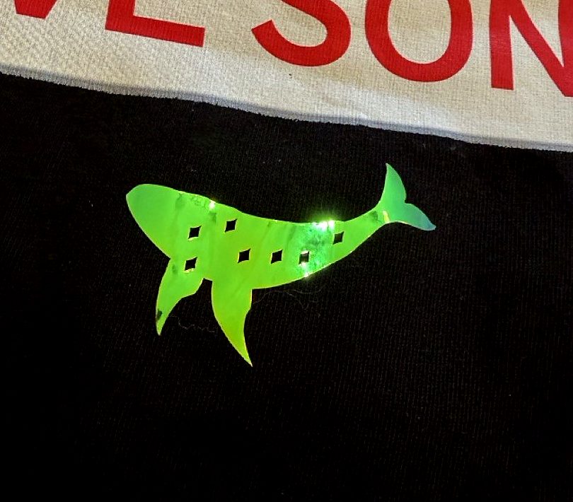 A green-gold holographic whale transfer pressed onto a black t-shirt below a printed slogan